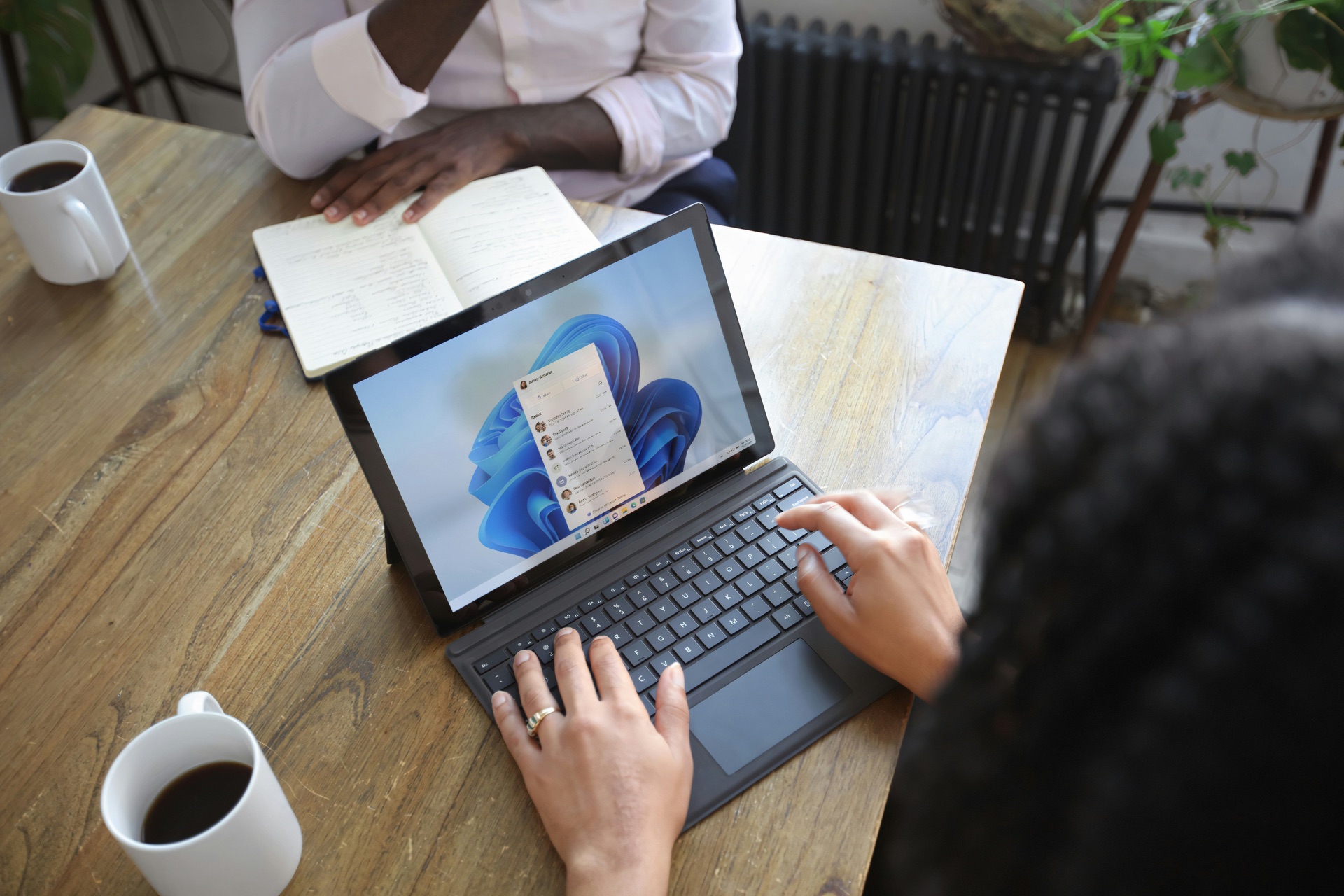 Two colleagues at a wooden table working on a Windows laptop showing a Microsoft 365 mail and Teams interface